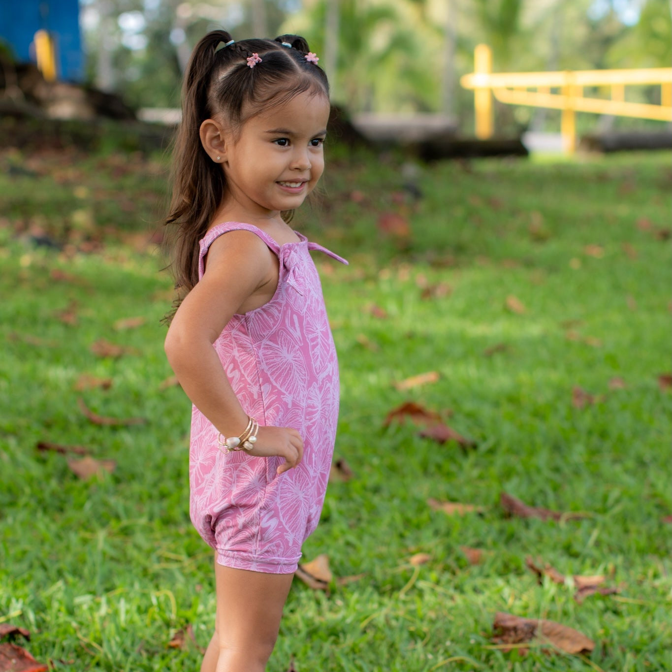 Young girl in a pink kalo overalls standing on grass with trees in the background