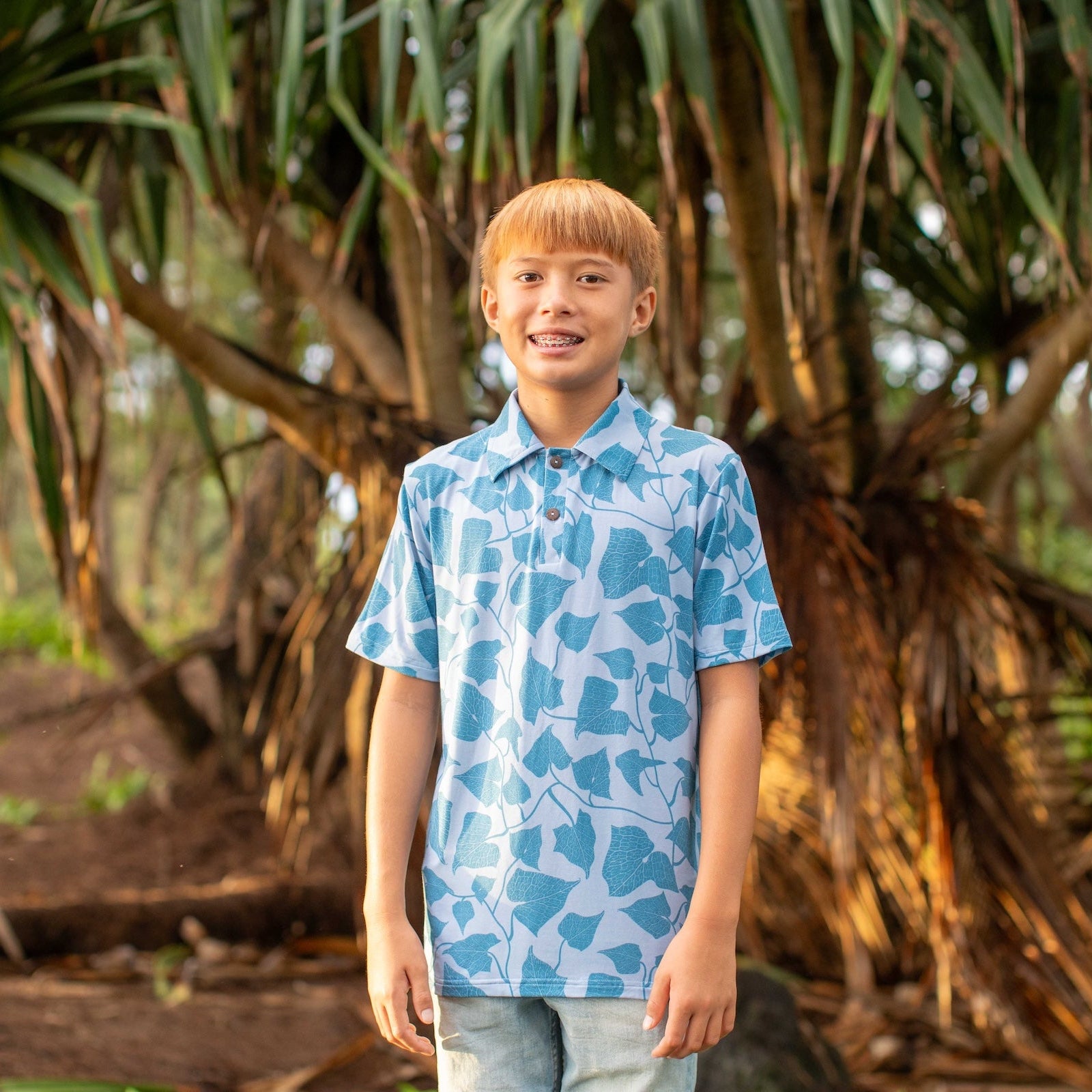 Young boy standing in front of a large tree with a blue patterned shirt.
