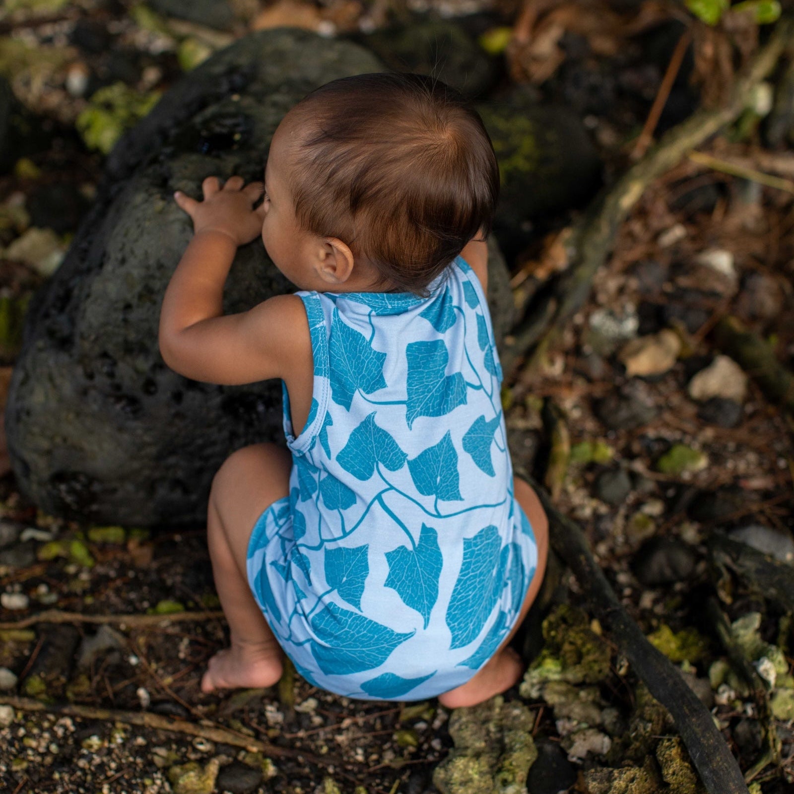 Baby in a blue leaf-patterned romper sitting on the ground outdoors.