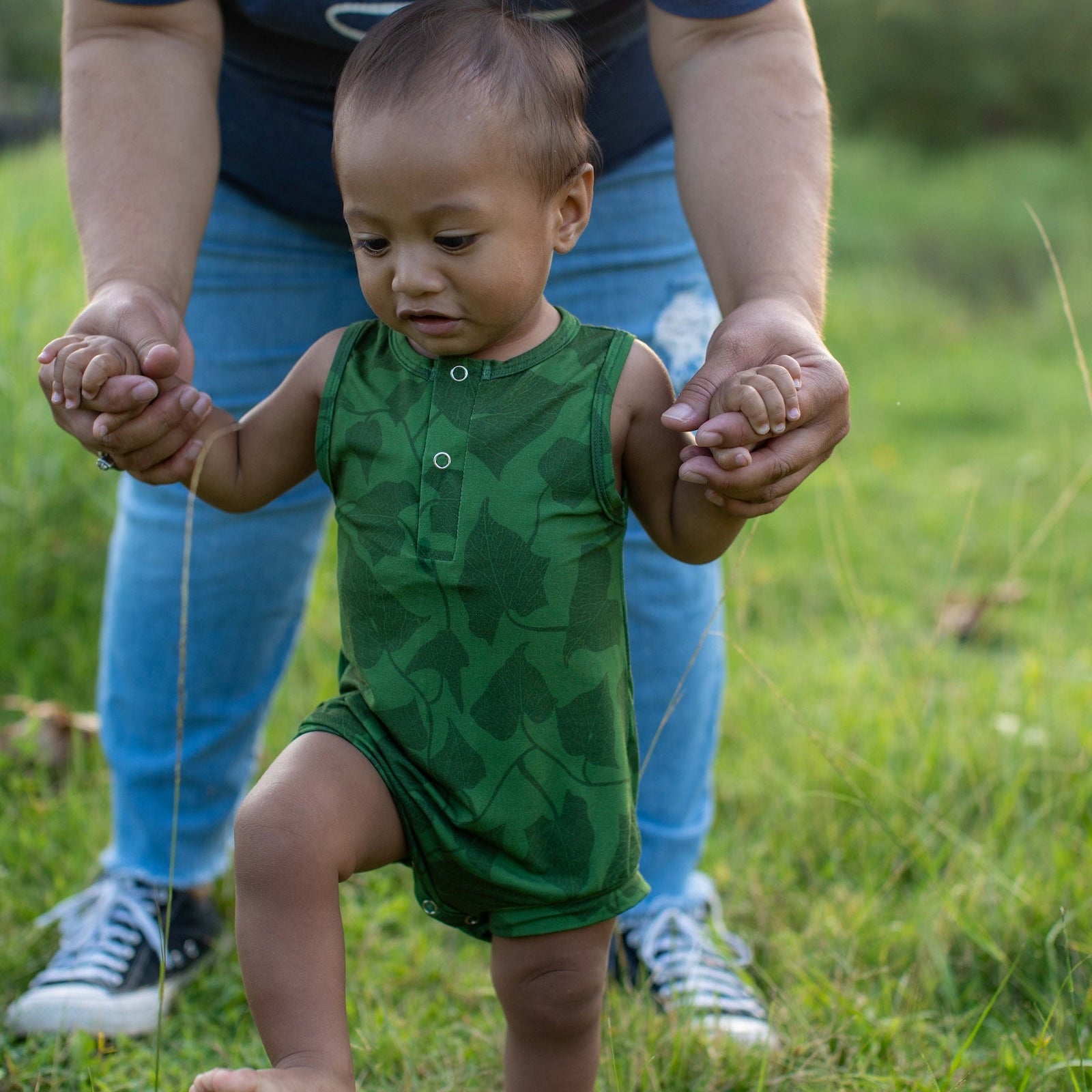 Child in a green outfit being held by an adult in a blue shirt and jeans outdoors.