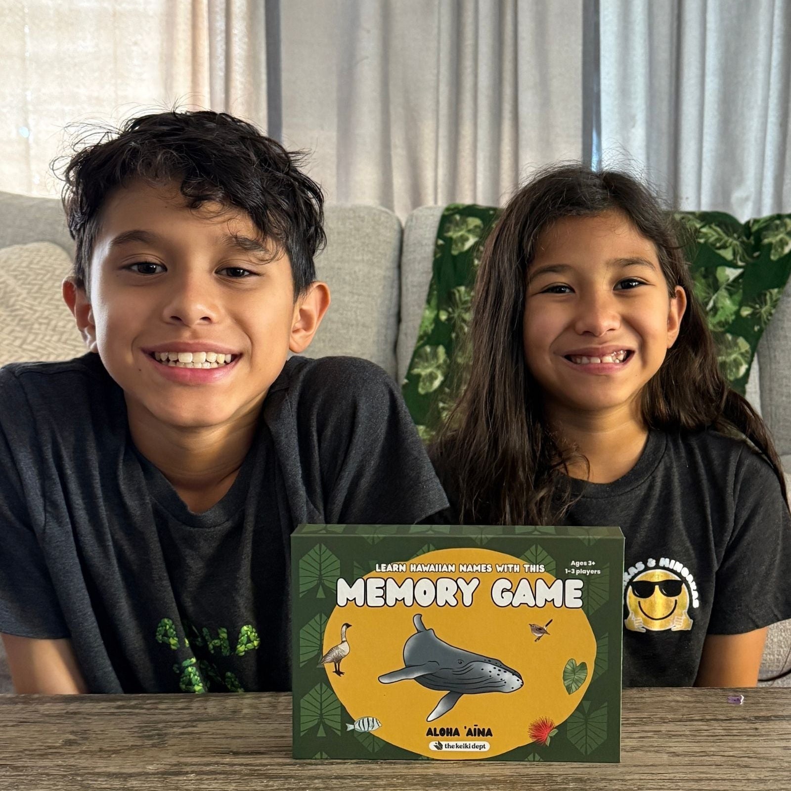Two children sitting behind a 'Memory Game' box on a wooden table.