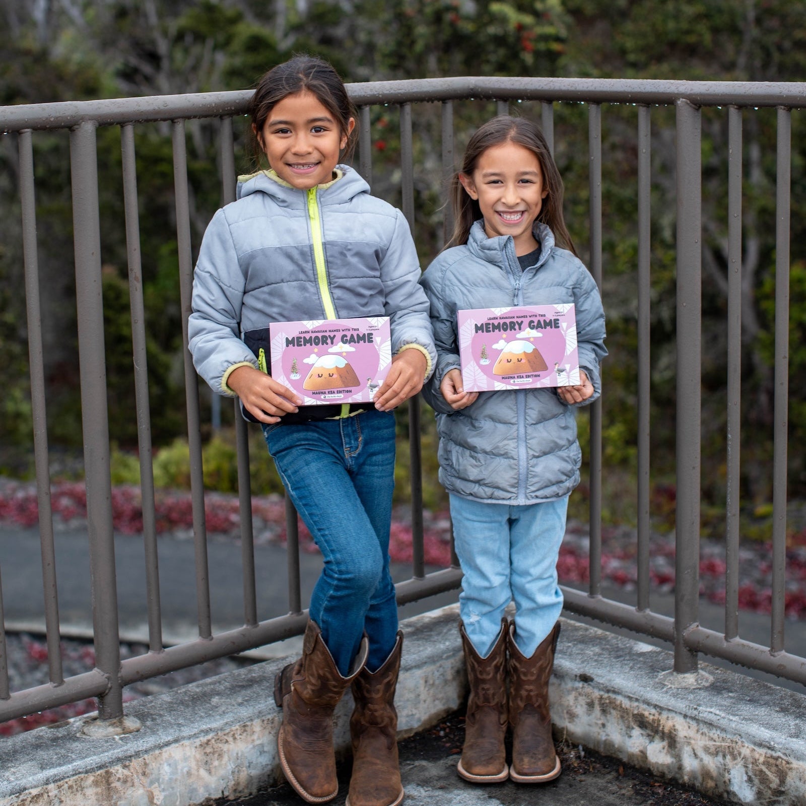 Two children holding pink game boxes with a metal railing and trees in the background.