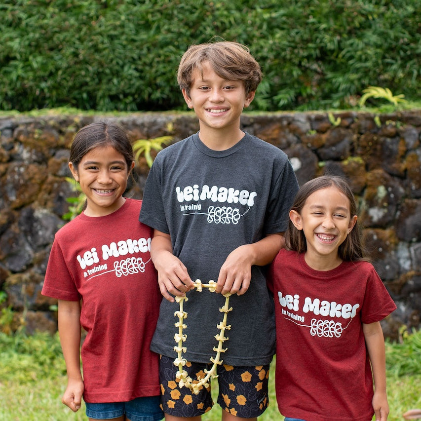 Three children wearing Lei Maker shirts standing outdoors with a stone wall and greenery in the background.