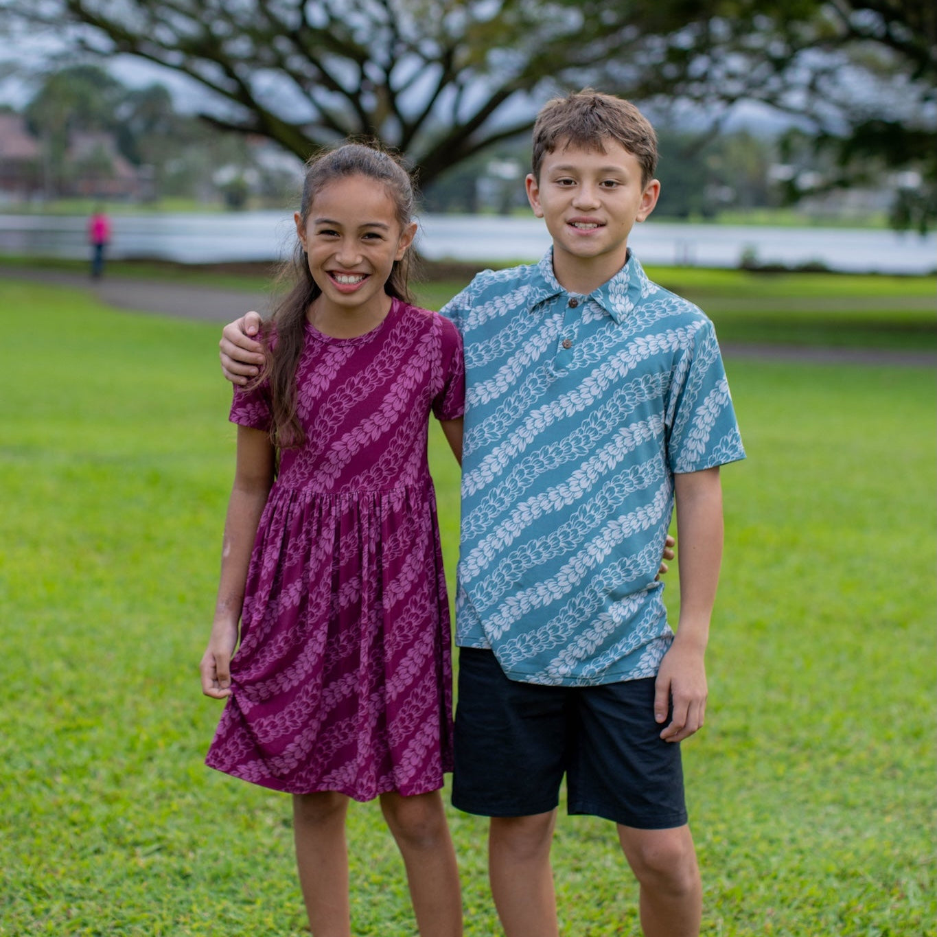 Two children standing on grass wearing pakalana strands dress and polo with trees in the background