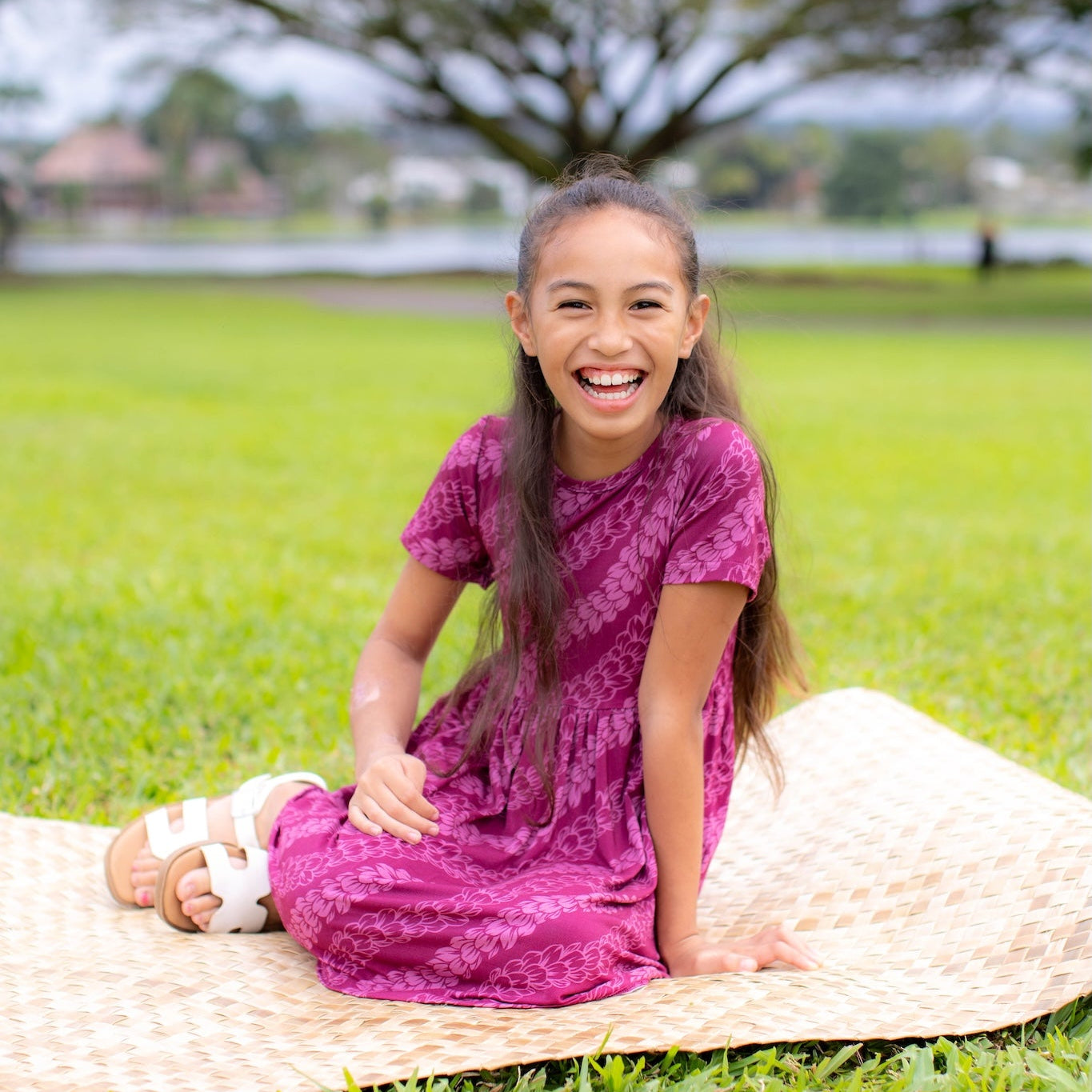 Young girl in a pink pakalana dress sitting on a woven mat in a park