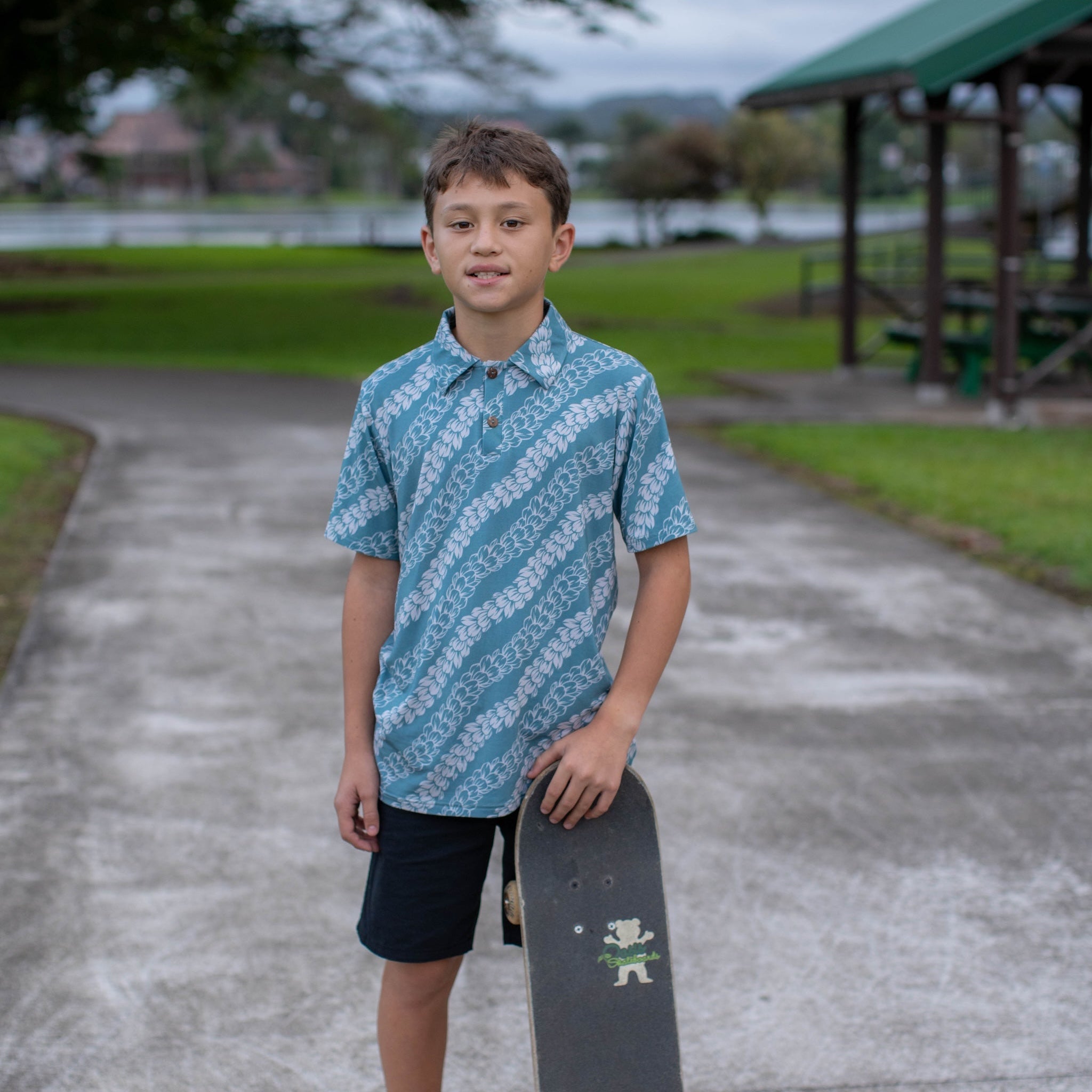 Young boy holding a skateboard in a park setting wearing a palaka strands polo shirt
