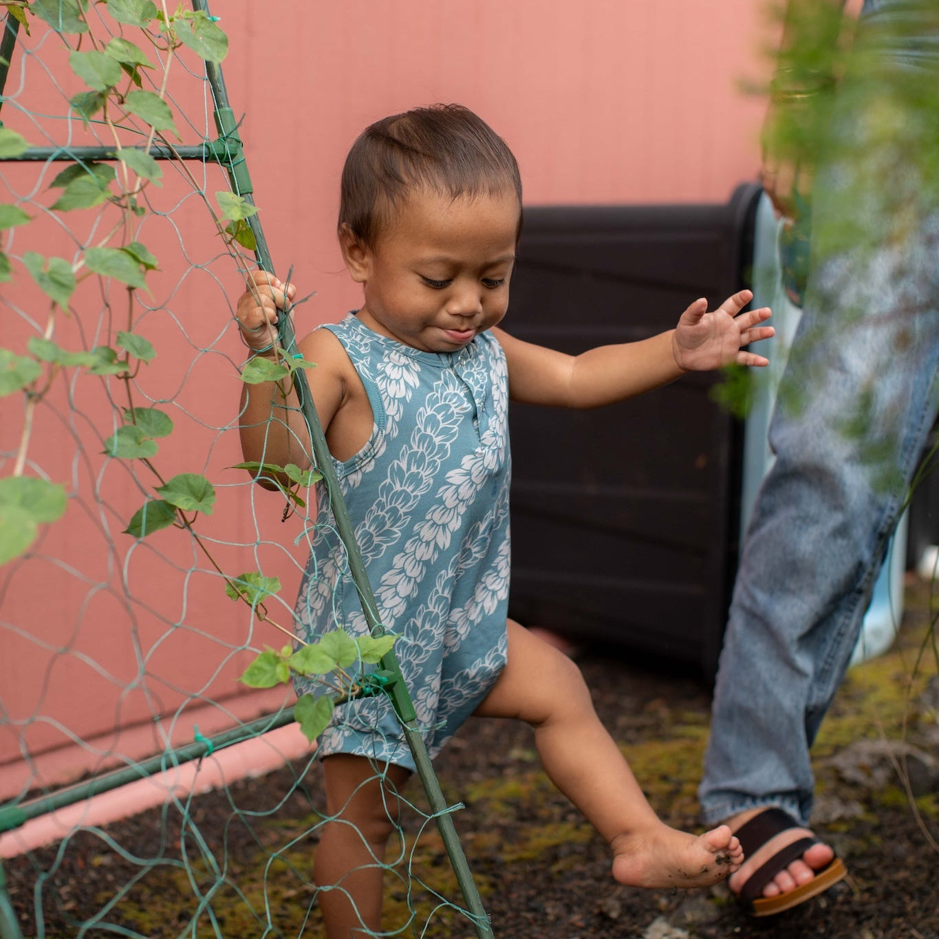 Child in a blue pakalana tank romper standing next to a plant trellis with a pink wall in the background.