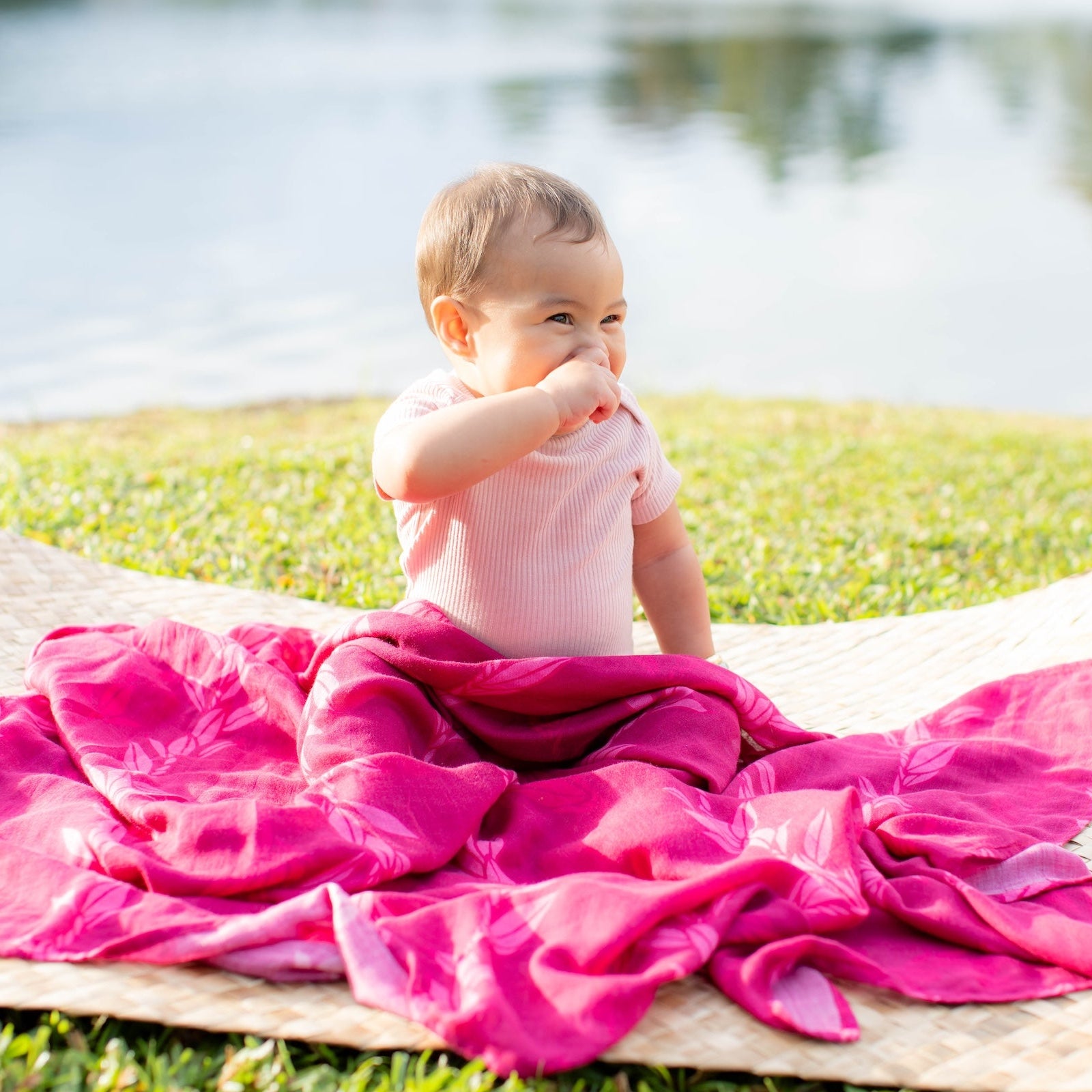 Baby sitting on a pink blanket outdoors by water